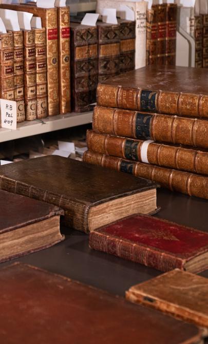 Shelf and table with dozens of old, leather-bound books, some with gold designs or writing on the spines