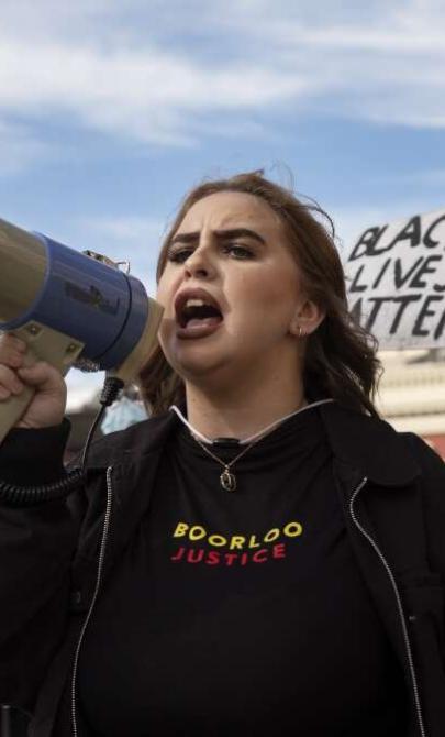 A person speaks into a megaphone during a protest rally, wearing a shirt with the text "Boorloo Justice." Behind them, others hold signs, including one that reads "Black Lives Matter." A yellow flag and buildings are visible in the background.