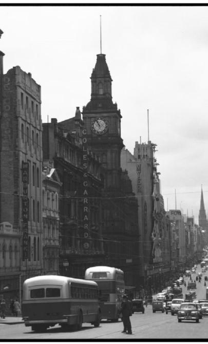 A black-and-white photograph showing Bourke Street, Melbourne, in the mid-20th century, with buses, cars, and pedestrians. The street is flanked by tall buildings with prominent signage, and a clock tower is visible.