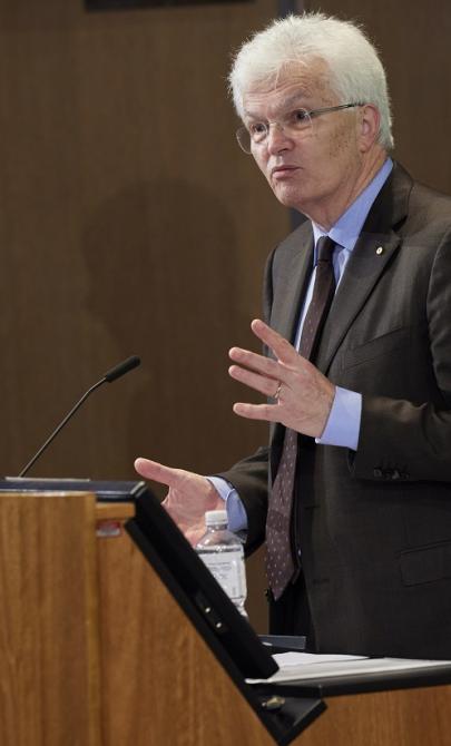 Professor Glyn Davis AC in a suit speaking at a podium and gesturing with his hands. Aboriginal and Australian flags are visible in the background.