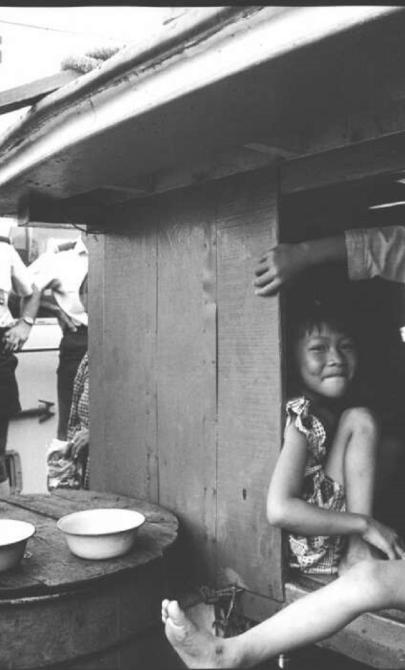 Three young girls smile and sit together inside a wooden boat cabin. A customs officer stands outside near a vessel labeled "Customs." Two bowls rest on a barrel near the cabin.