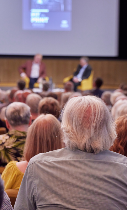 Looking over people in a crowded theatre towards two men sitting on the stage chatting