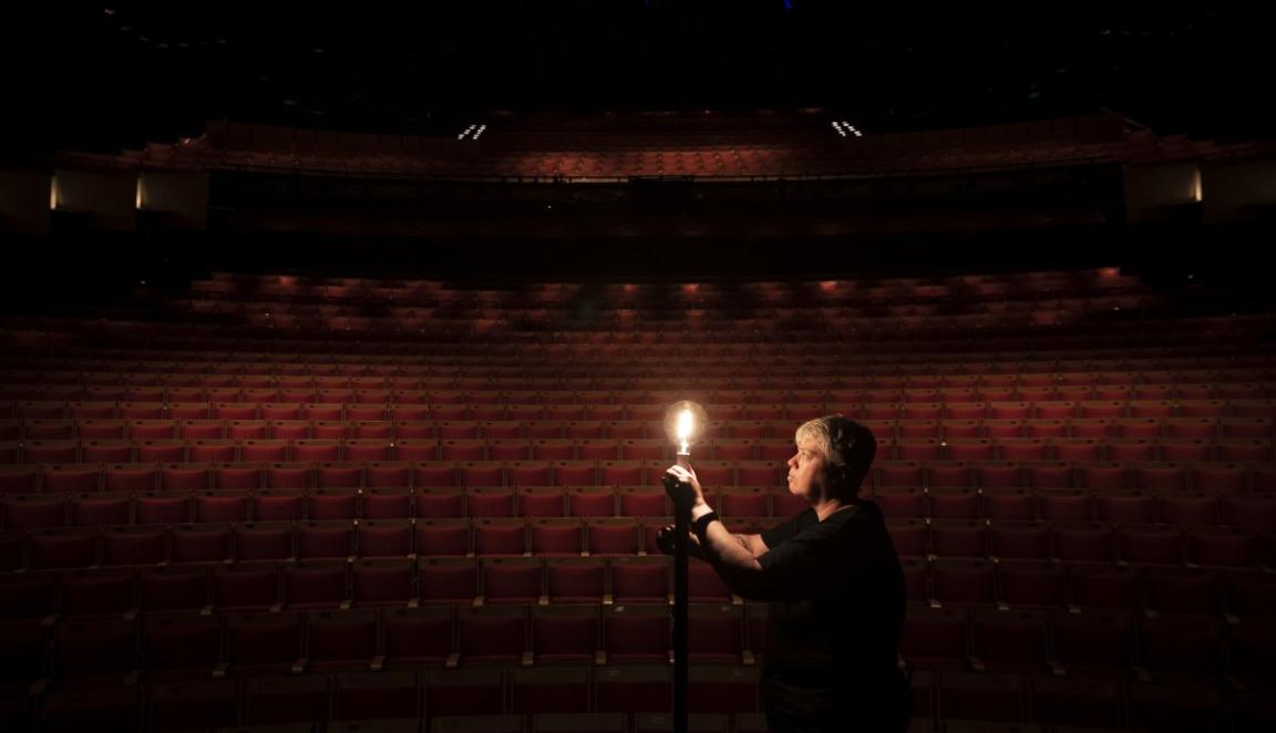 A person stands in an empty darkened theatre, illuminated by a single light bulb.
