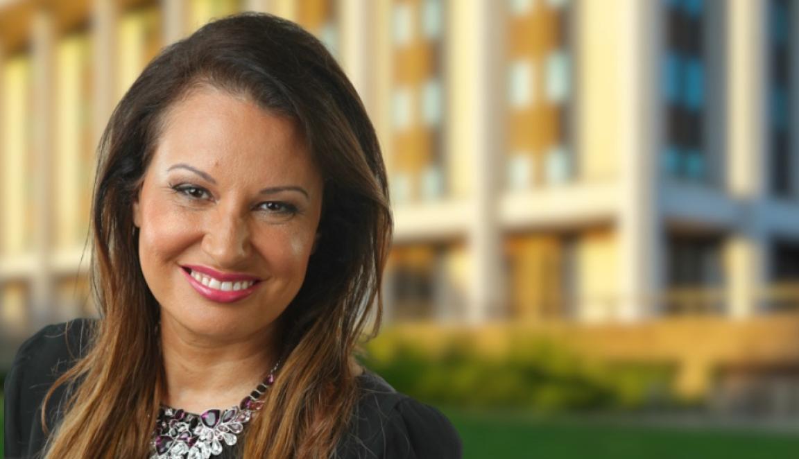 Eualayai/Gamillaroi woman with long brown hair wearing a black top and statement necklace smiling and standing in front of the National Library building