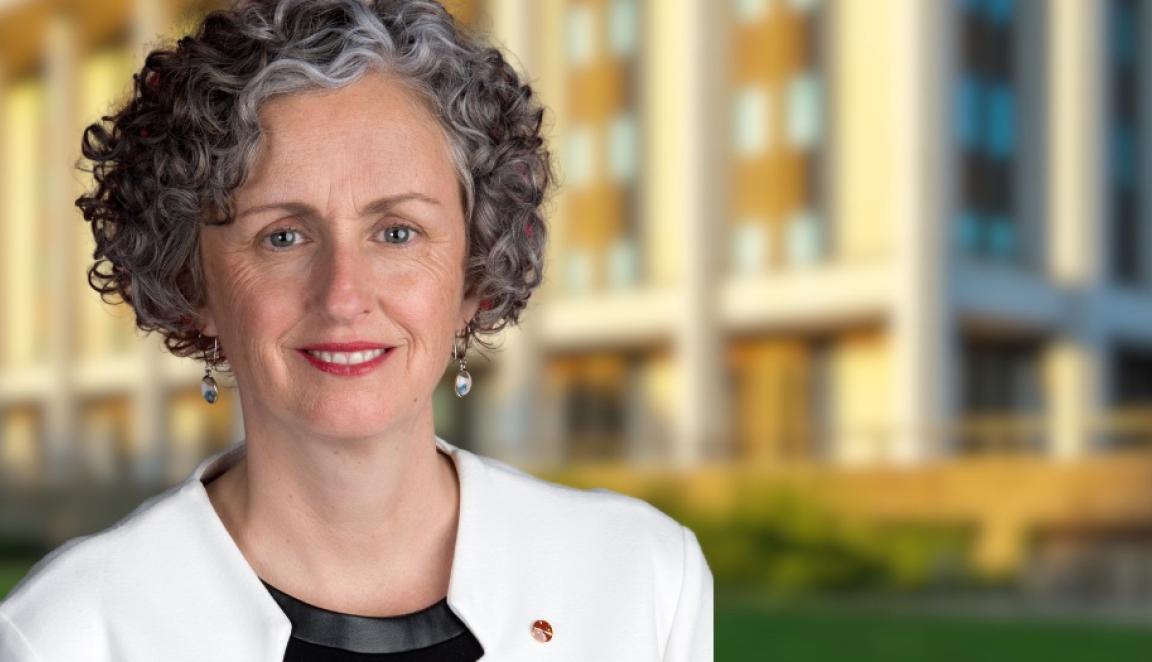 Woman with short, curly black and grey hair wearing a black top and white blazer standing in front of the National Library building