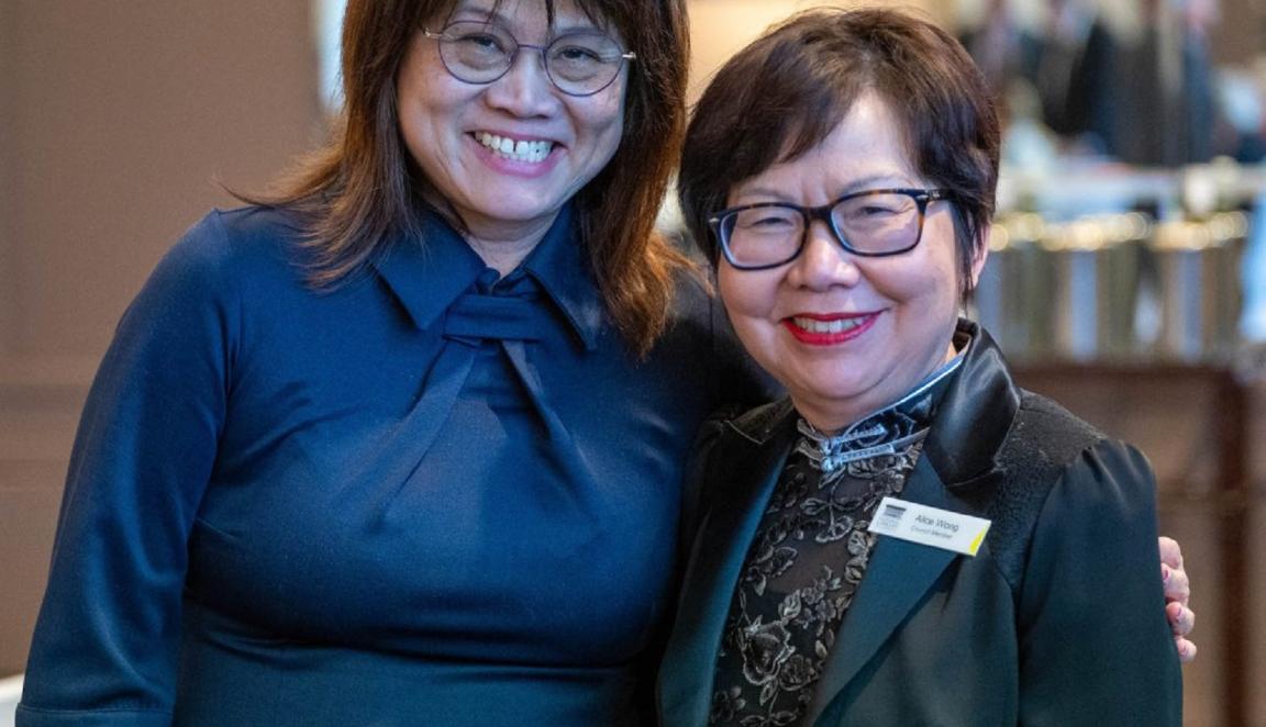 Two people standing next to each other, smiling. One has shoulder-length dark brown hair and wears glasses and a dark blue dress with a bow detail at the neckline. The other has short dark hair, also wearing glasses, and a black jacket with a floral pattern on the lapel, along with a name tag.