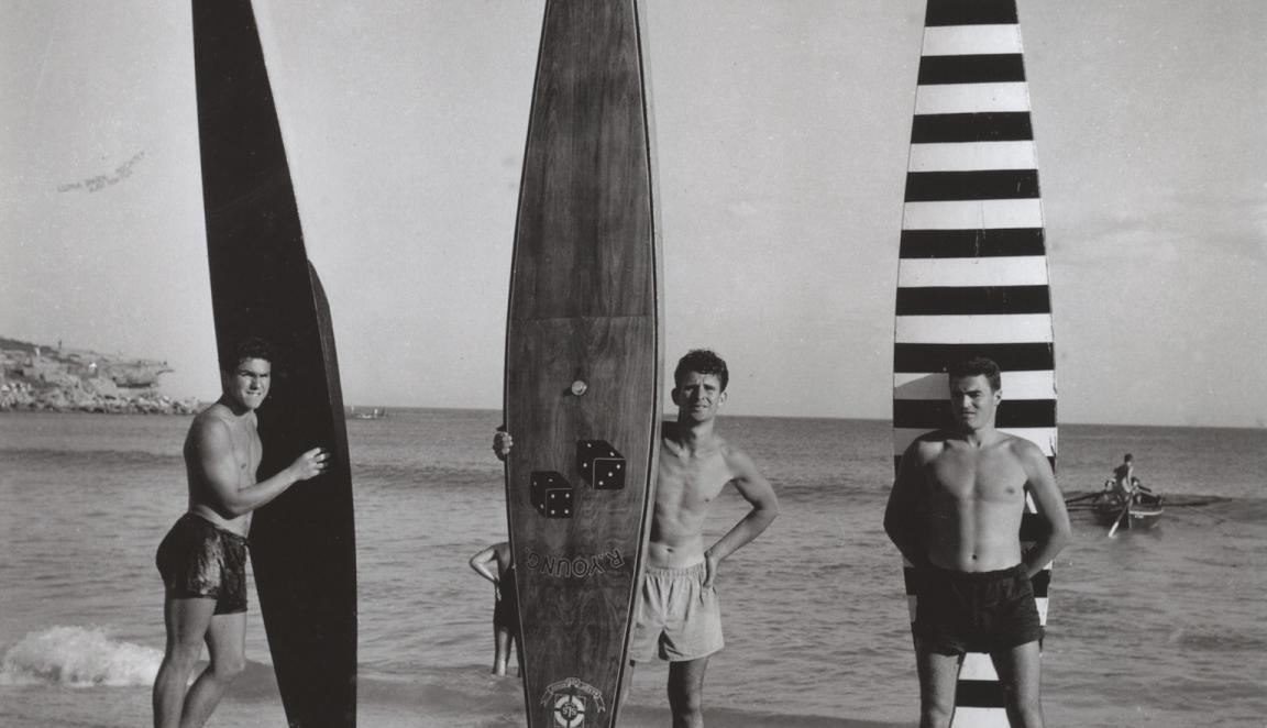 Black and white photo showing three men in swimwear holding up long boards on the sand at a beach