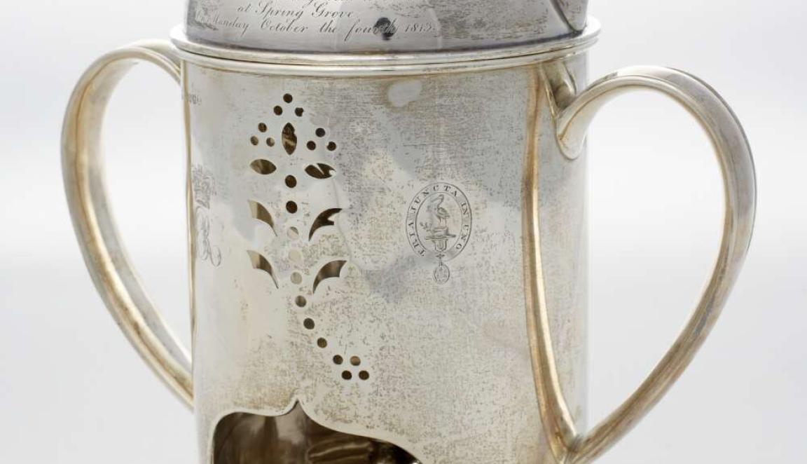 A silver polished kettle with ornate cutouts and inscription and printed crest. It has a tealight underneath.