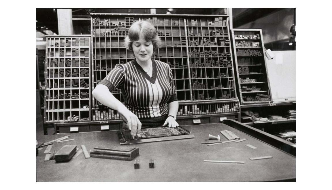 A black and white image of a woman at a desk setting type face into a frame. Behind her is a large rack full of letters