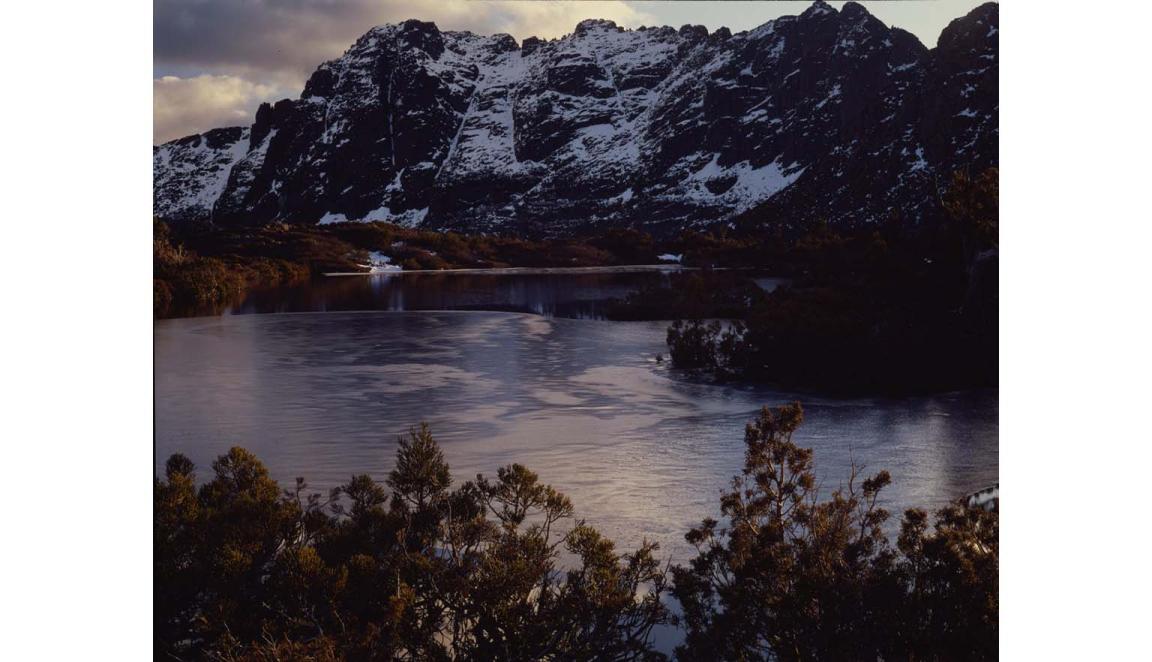 A landscape of a mountain lake scene. The craggy mountains are dusted with snow. The lake is surrounded by trees with yellow leaves.