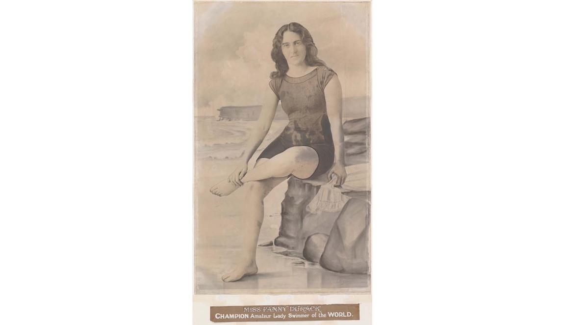 A sepia toned photograph of a woman sitting on a rock. She is wearing an old fashioned swim suit. She is sitting crossed legged. Beneath her is a caption saying 'MISS FANNY DURACK' Champion Amateur Lady Swimmer of the World'