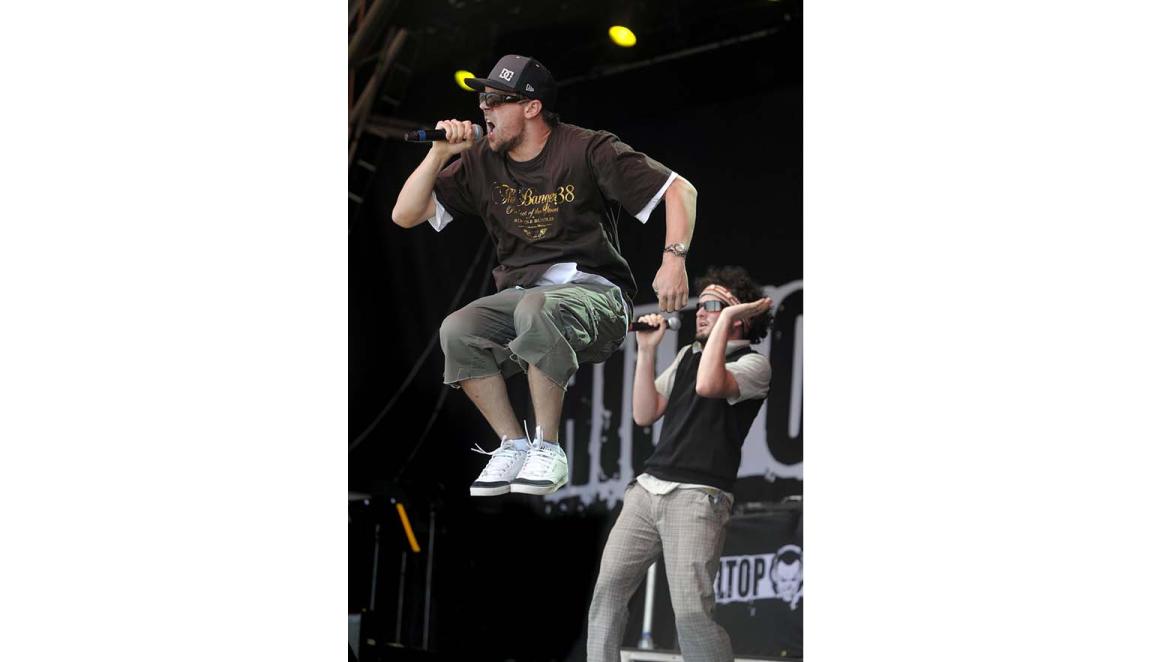 A man in shorts, tshirt, cap and sunglasses is depicted jumping in mid-air, singing into a microphone. Another musician stands behind him.