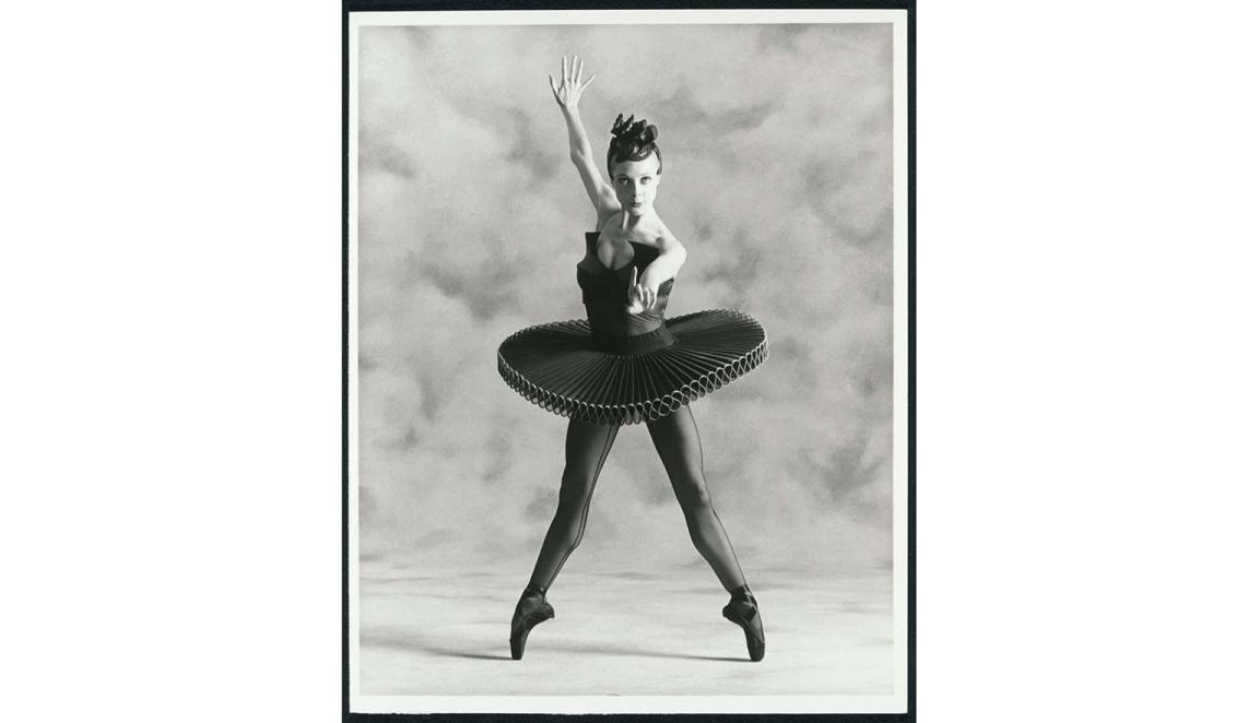 Ballerina Justine Summers strikes a pose and stares at the camera. She is standing en point wearing a black tutu. She is standing infront of a textured curtain.
