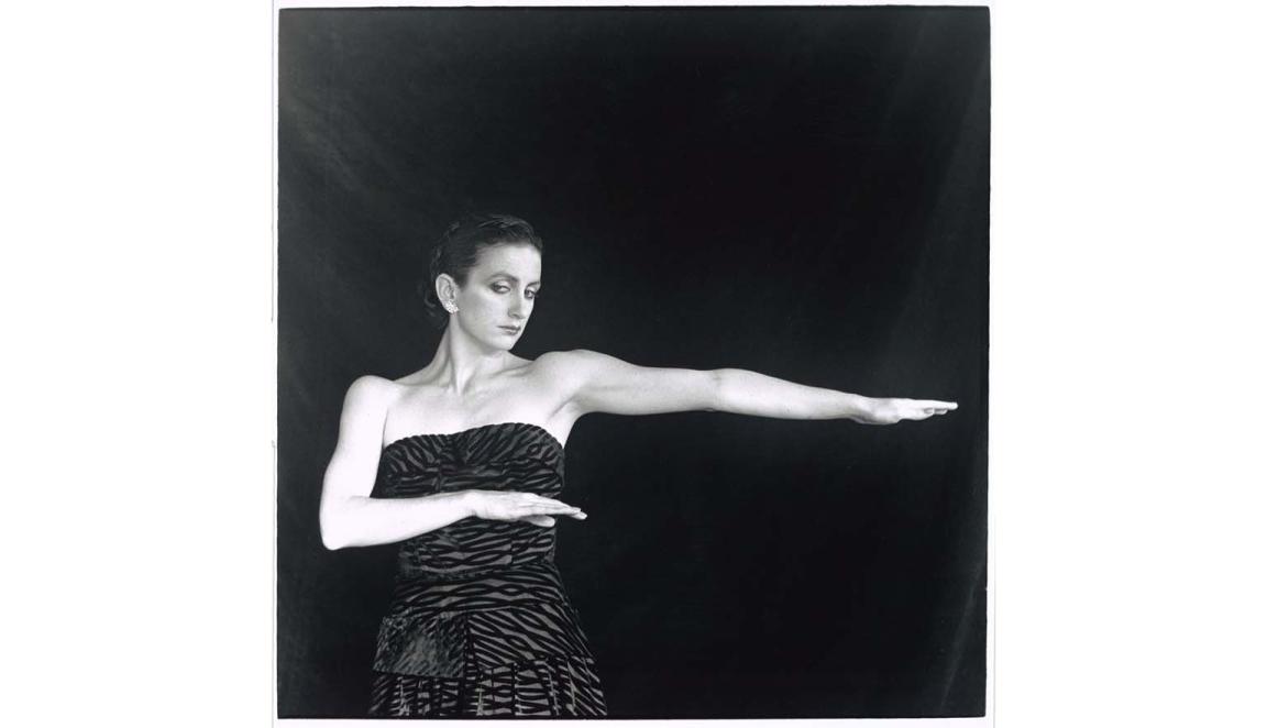 Ballerina Meryl Tankard strikes a pose. She is wearing a zebra print-esque dress. She is standing in front of a black curtain.
