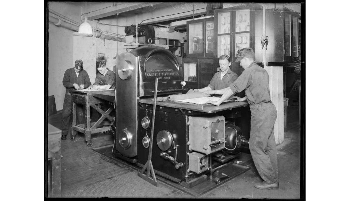 A black and white image of a group of men standing around a large industrial press.