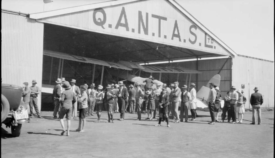 Black and white photo of a crowd of people examining a biplane in a QANTAS hanger