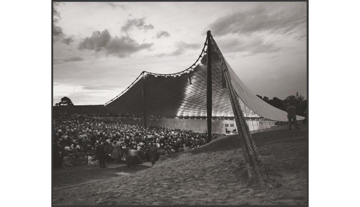 A sepia photograph of the Sydney Myer Music Bowl. A large trianglular sail like building with a stage. A large crowd are gathered watching the stage.