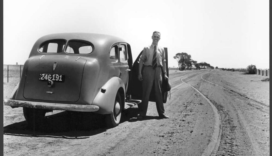 Black and white image of a man in a suit standing next to a car on a dirt road. 