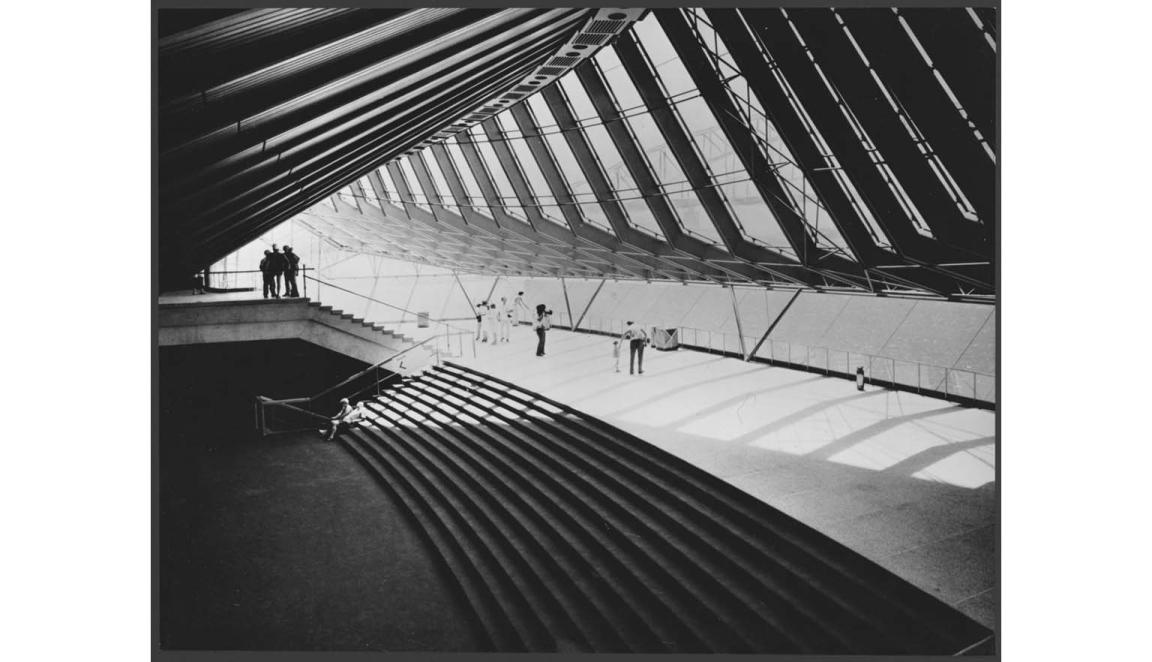 A black and white photograph of the interior of the Sydney Opera House. The windows give an effect of being in a large rib cage. There is a large sweeping staircase taking up most of the image