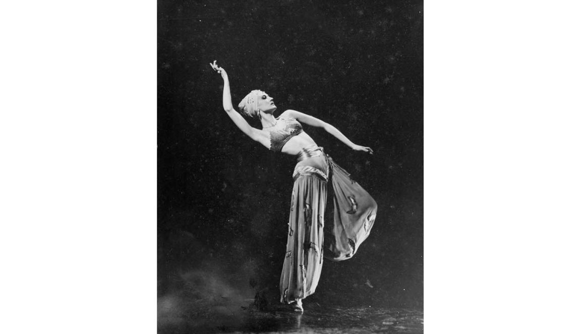 A woman in a headwrap and long pants is on stage; she leans backward with an arm raised above her head