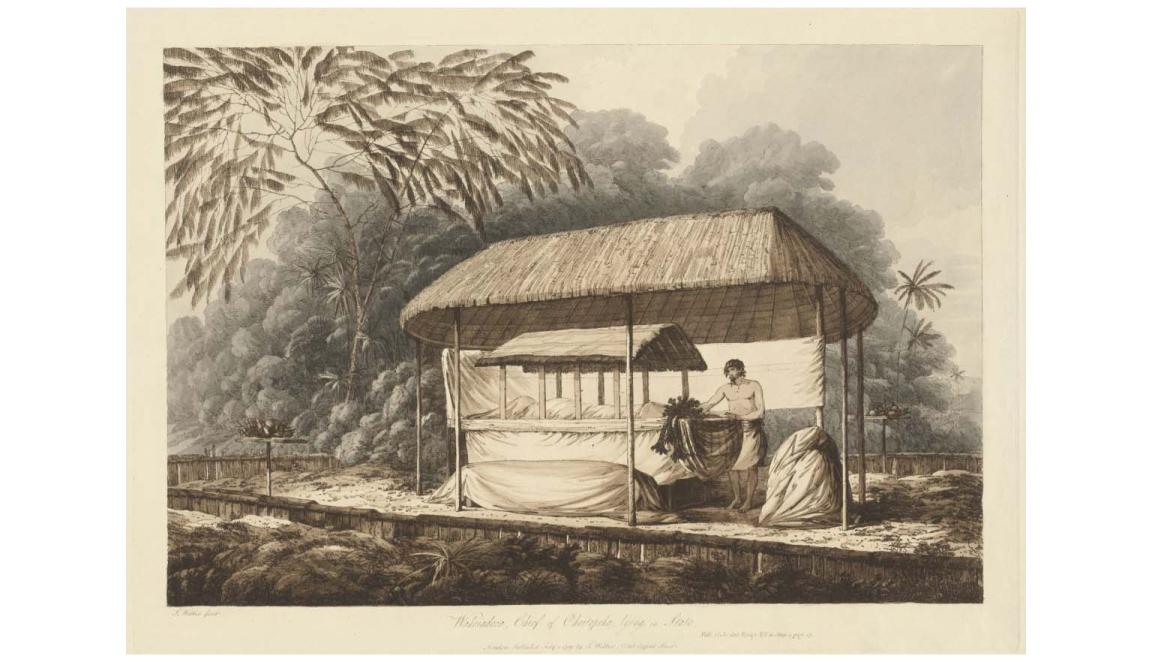 A man stands next to a ceremonial alter. The alter has a thatched roof. It sits inside a larger shelter also with a thatched room. On the alter is a body shrouded in cloth. Behind the shelter is a large thicket of trees.
