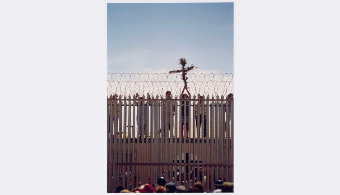 A wooden cross wrapped with foliage is held high by hands reaching through vertical metal bars topped with barbed wire.