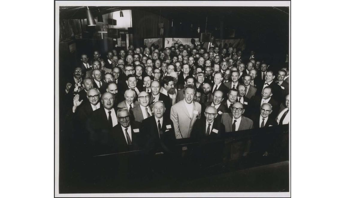 A black and white photograph of a large group of men, mainly older, standing in a large room. All the men are wearing dark suits and ties except one man in the centre of the photo. He is wearing a white suite with a white turtleneck sweater. Many of the men are wearing glasses. Some are waving at the camera and almost all are smiling broadly.