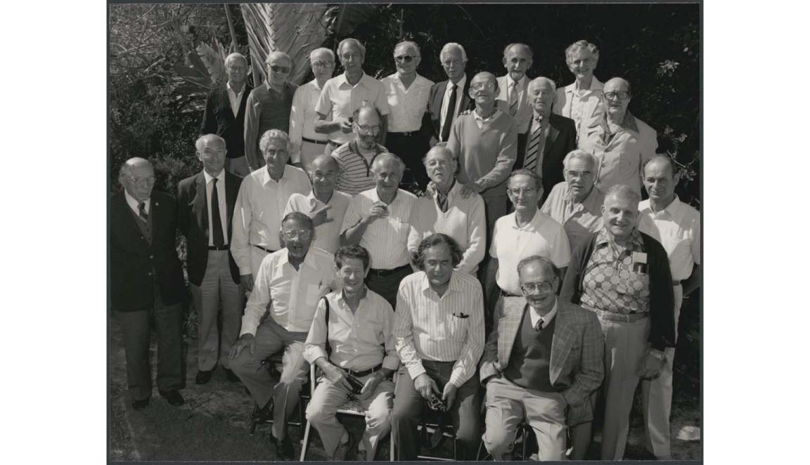 A black and white photograph of a large group of older men, posed for a group photo.  Most of the men are smiling at the camera. Some men are sitting in the front rows while others stand. They are smiling at the camera.