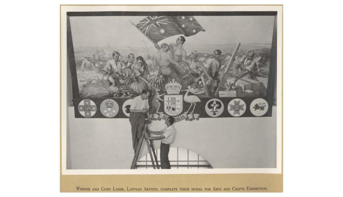 Two men, one standing on a ladder and one holding it steady, looking up at a mural they painted depicting several men holding up an Australian flag