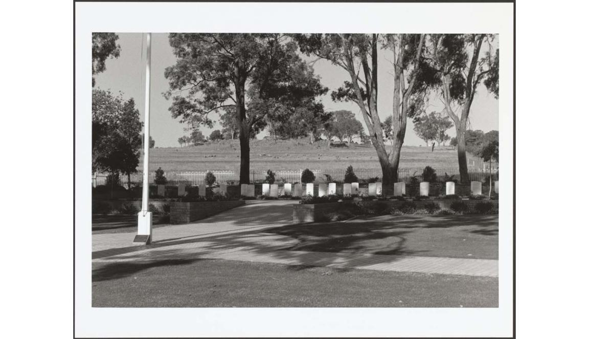 A black and white photograph of a memorial area. There are a row of 23 white headstones along the middle of the image. They are in the shade of large gumtrees. There is a white flagpole on the left of the image. In the background there is a grassy hill.
