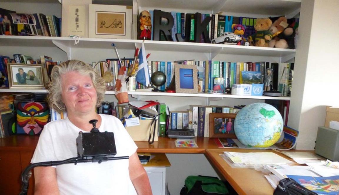 A photograph of a smiling woman seated in a wheelchair in front of a large bookcase and desk. She is wearing a white tshirt. The wheelchair has a control stick which sits just below her chin.