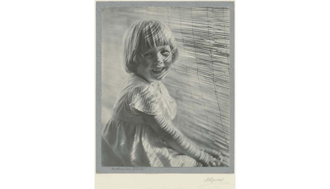 Black and white photo of a young girl smiling at the camera and sitting in front of a bamboo blind with sunlight lines across her face. 'The Bamboo Blind' is handwritten on the bottome left and 'H Cazneeaux' on the bottom right