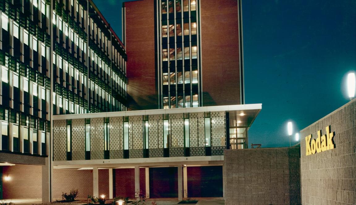 Coloured photo of the Kodak factory, a modernist architectural building, at night, with the lights inside the building glowing