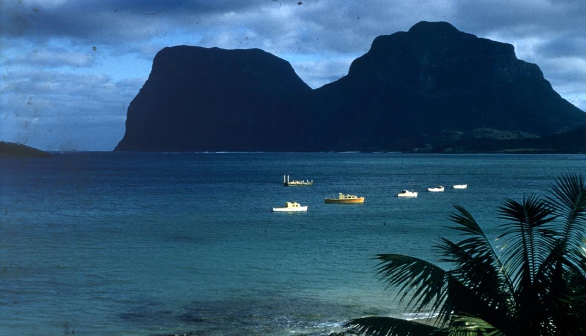 A colour photograph of the Lagoon in Lord Howe Island, two mountain peaks in the background, blue flat sea and five small boats are visible in the foreground.