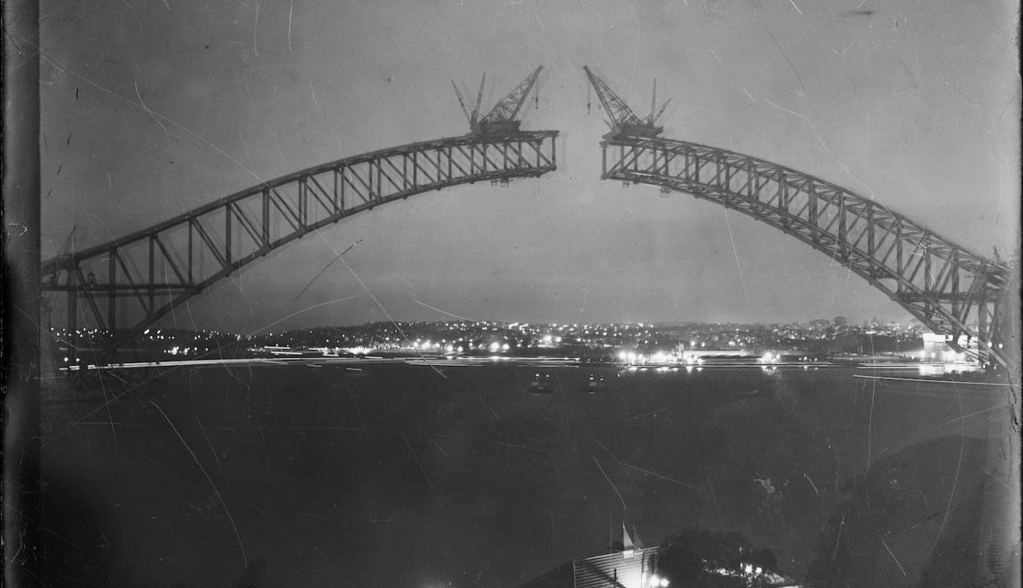 Black and white photo of metal being lowed by crane to complete the top of the Sydney Harbour Bridge