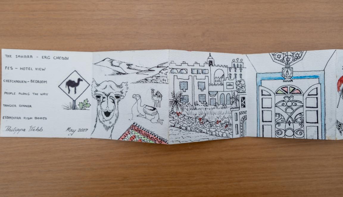Fold-out travel diary open to three pages with black and white illustrations of camels, Moroccan buildings and a door