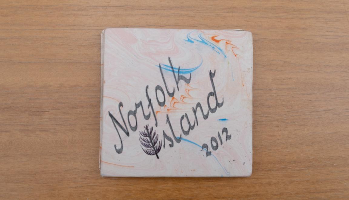 Travel diary cover with an orange, white and blue swirl design and large black writing reading 'Norfolk Island 2012' with the 'I' of 'Island' being a tree