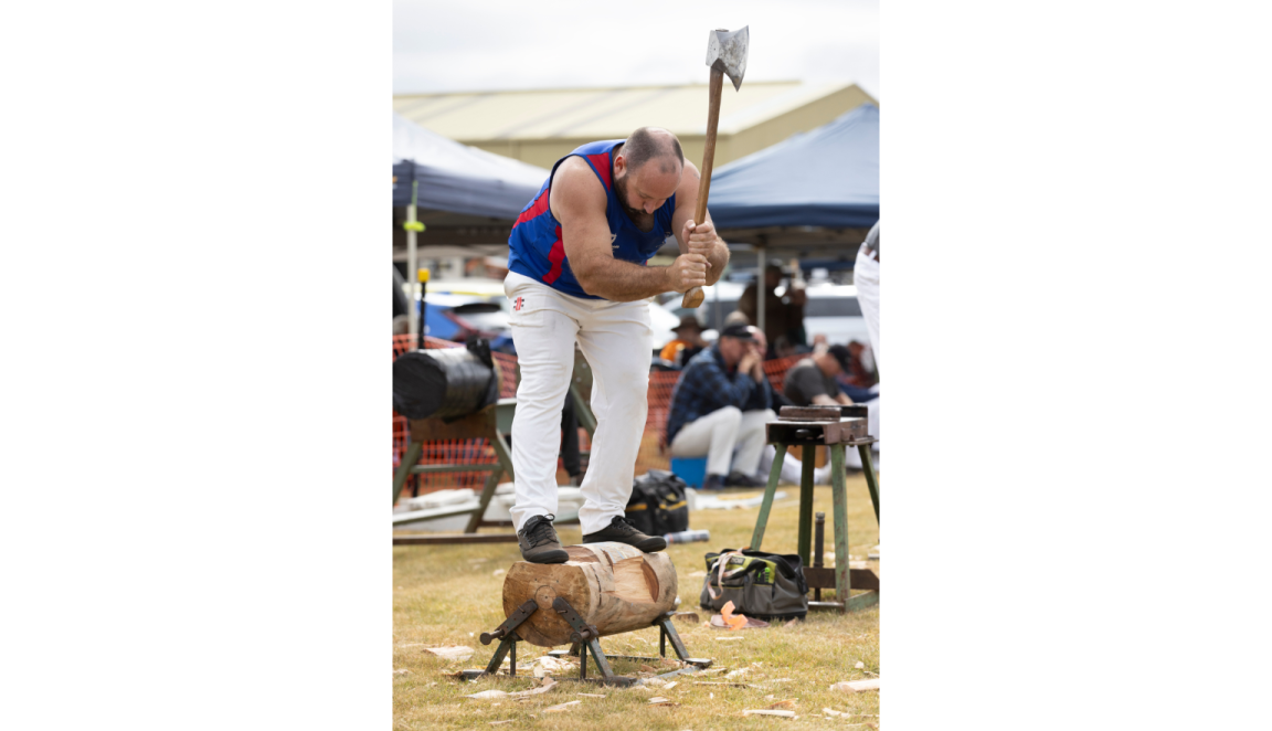 Man standing on a block of wood held up slightly above the ground and swinging an axe up to chop it in half
