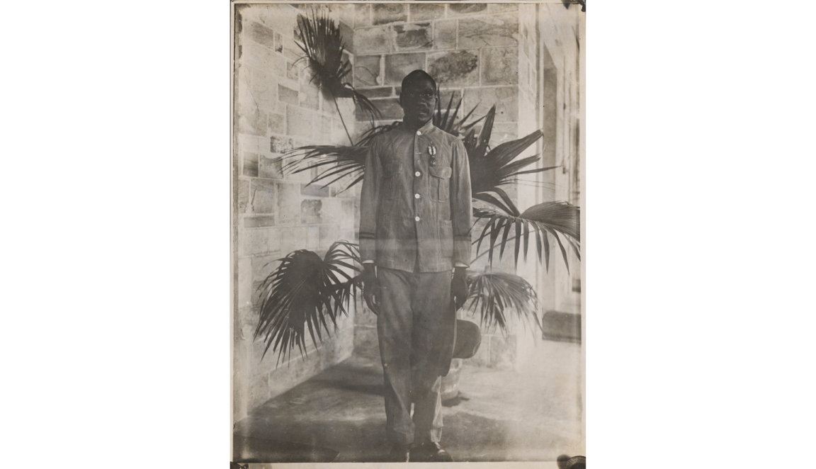 A photograph of an Indigenous man standing on the porch of a large house. Directly behind him is a large palm plant. He is staring at the camera and is standing slightly uncomfortably. He is wearing a military style tunic. Pinned to his left breast above the pocket is a medal. The ribbon is vertically striped.