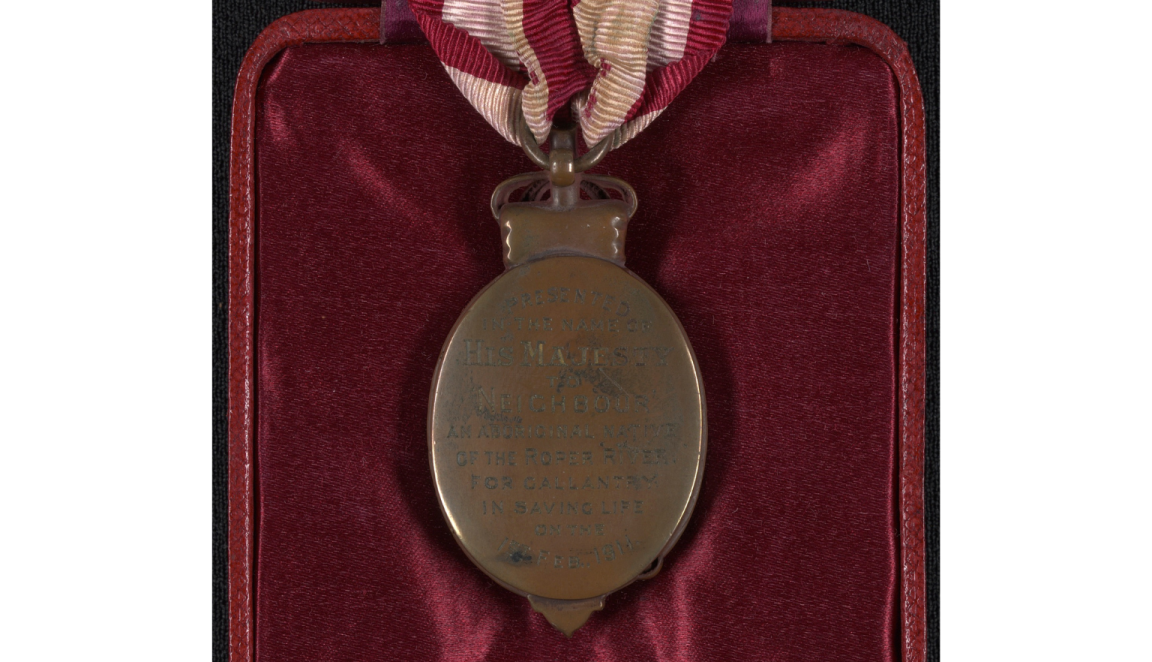 The reverse of an oval shaped bronze medal held by a red and white striped ribbon. On the back of the medal are the words 'Presented in the name of His Majesty to Neighbour, an Aboriginal native of the Roper River for gallantry in saving life on the 1st Feb 1911'