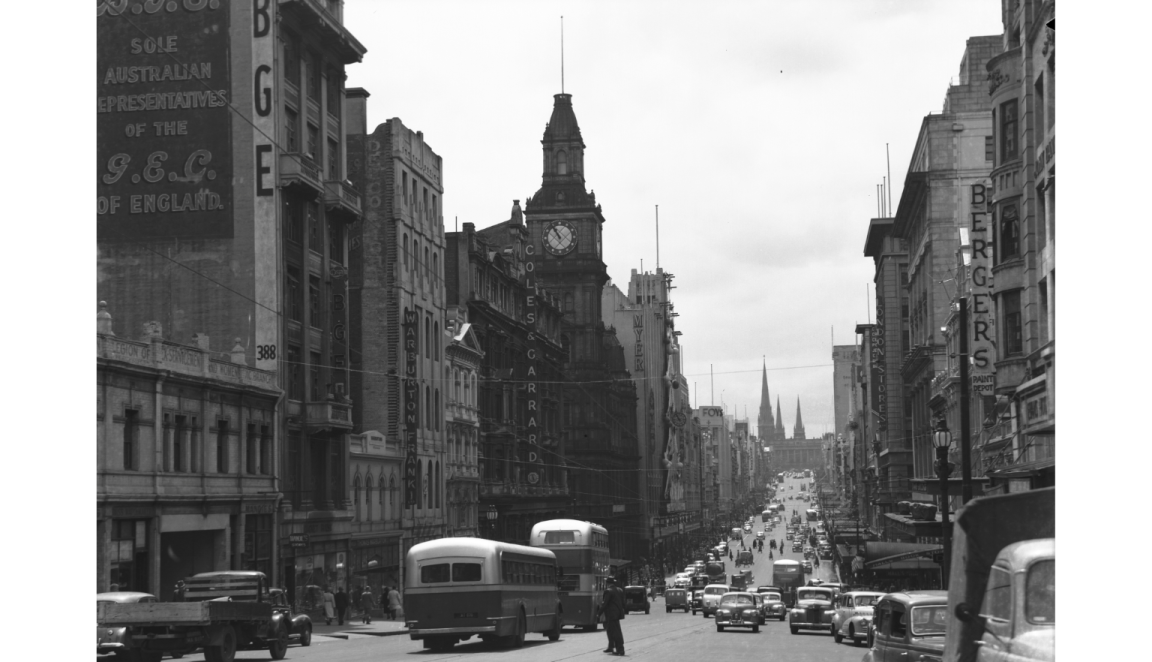 A black-and-white photograph showing Bourke Street, Melbourne, in the mid-20th century, with buses, cars, and pedestrians. The street is flanked by tall buildings with prominent signage, and a clock tower is visible.
