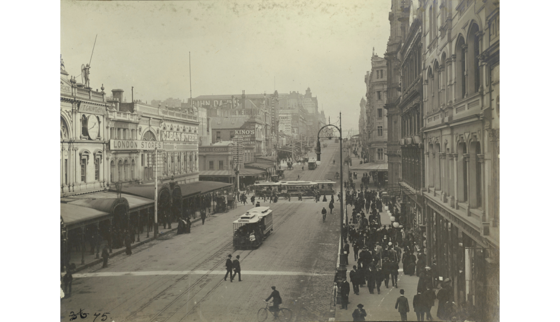A black-and-white photograph of early 20th-century Bourke Street, Melbourne, with trams, pedestrians, and people on bicycles. The street is lined with ornate building facades, and a crowd walks along the footpath.