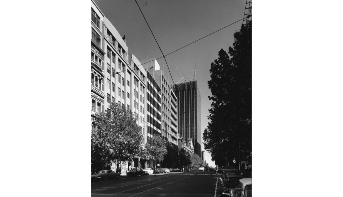 A black-and-white photograph of Bourke Street, Melbourne, showing mid-century modern buildings, with the AMP-St. James building under construction. Cars are parked along the street, with trees lining the road.