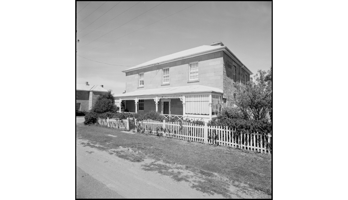 Black and white photo of a wo-storey sandstone house with a white picket fence
