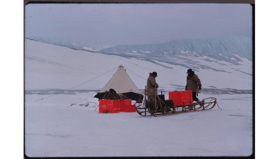 Two men in brown snow coats facing each other behind a sled and in front of a pitched tent, surrounded by snow