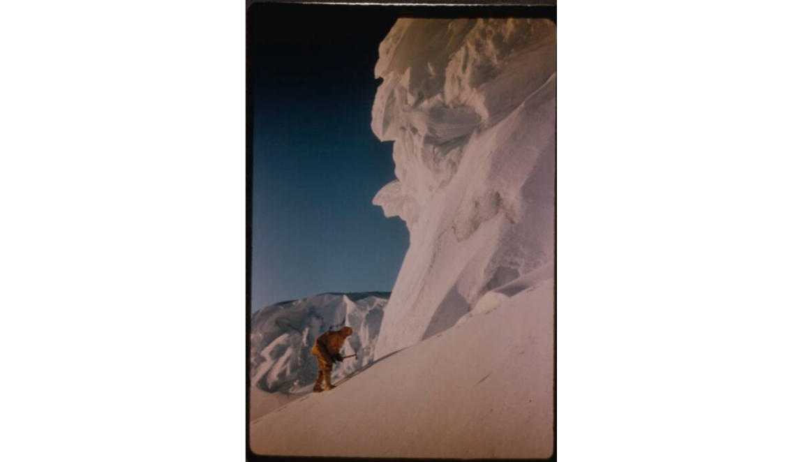 Man in yellow snow gear, bending down in front of a large cliff of ice and snow