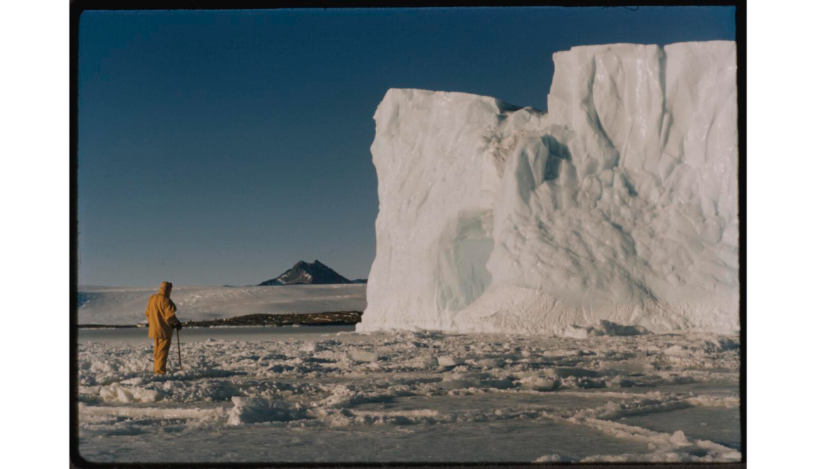 Man in yellow snow gear, facing a large cliff of ice and snow