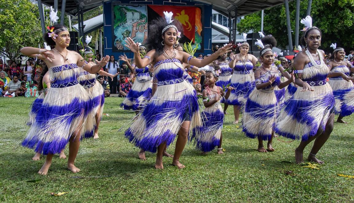 Several women dancing in rows with their arms out, wearing tassled purple and white dresses