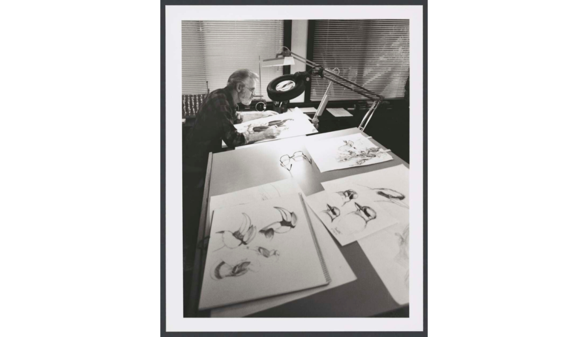 A black-and-white photo of an older man with glasses, seated at a desk and sketching birds. Several bird drawings are spread across the desk, and a magnifying lamp illuminates his work area. The scene suggests a dedicated artist or illustrator in their studio.