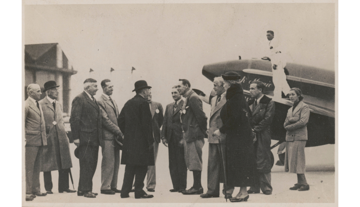 Black and white photo of a group of people standing on an airfield next to a plane.